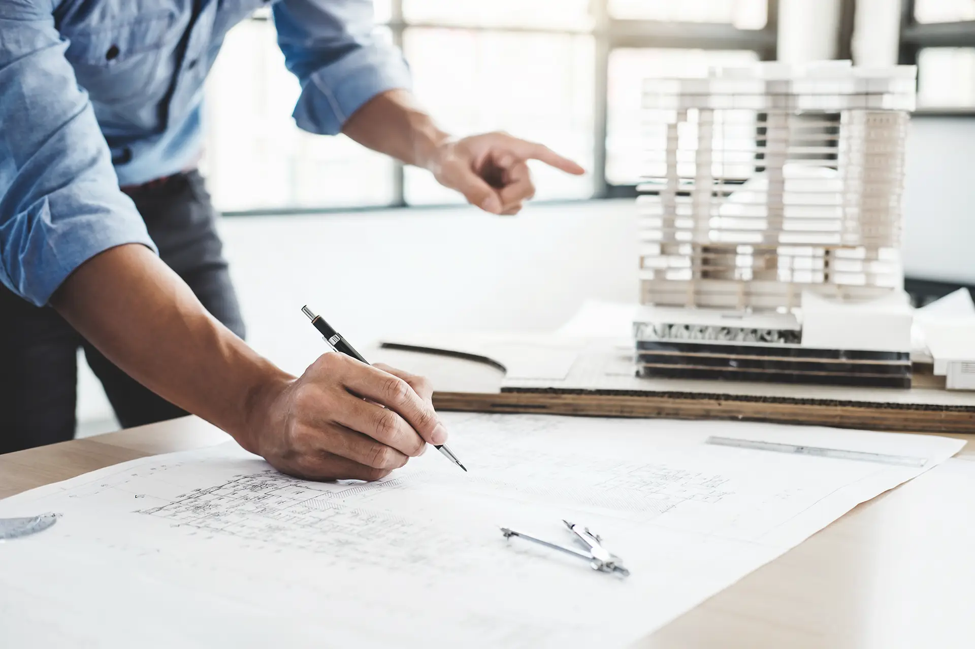 Mãos desenhando uma planta arquitetônica ao lado de uma maquete